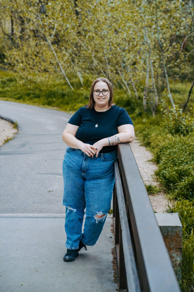 A woman with glasses and a tattoo on her right arm leans on a wooden railing along a paved path in a green, tree-filled park, smiling at the camera.