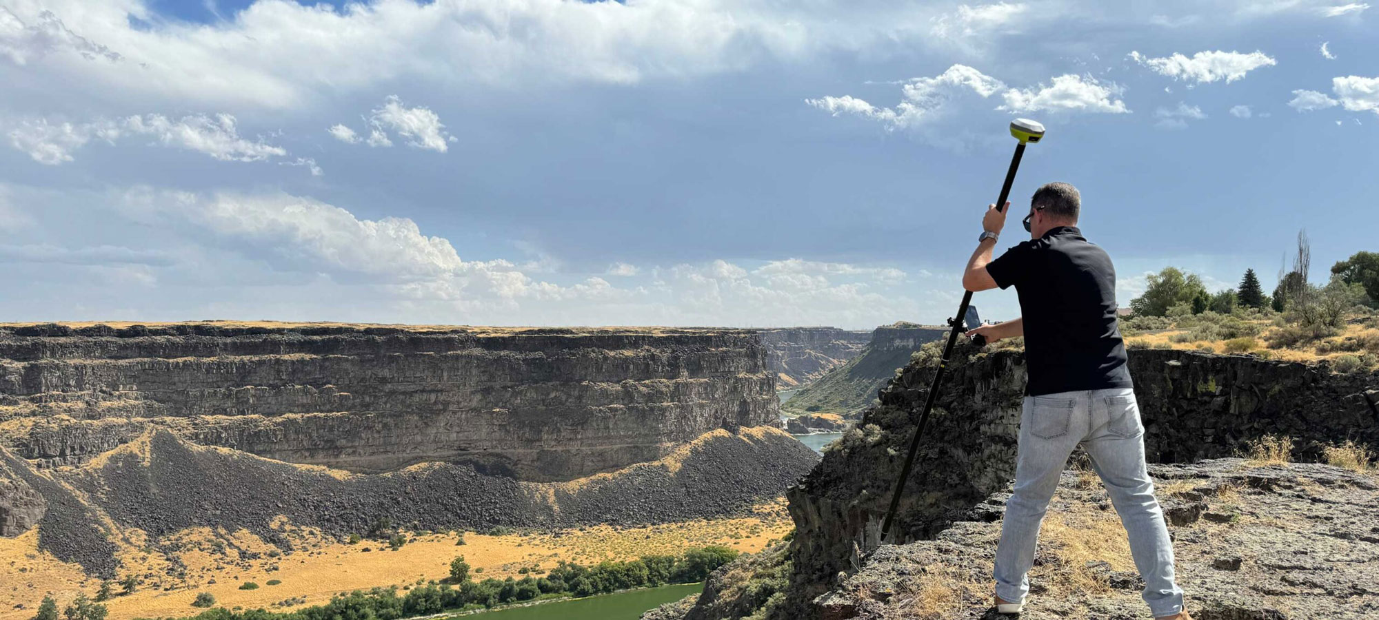 A person stands on a rocky cliff holding surveying equipment, overlooking a deep canyon with layered rock formations and river below, under a partly cloudy sky.